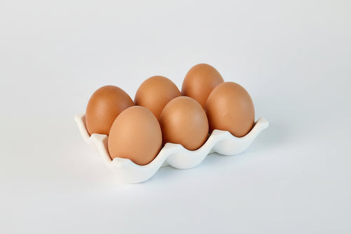 Six brown eggs in a white ceramic carton on a light gray background